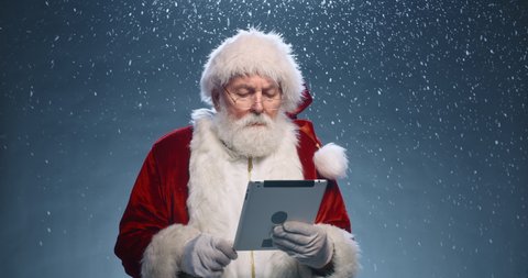 Santa Claus surfing internet on his tablet, then looking at camera nad winking, isolated over snowy blue background - christmas spirit concept close up 4k footage