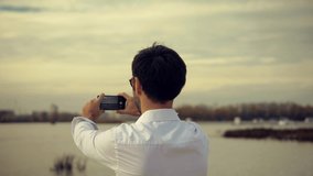 Man Takes Pictures Smartphone On City Beach. Handsome Male Taking Mobile Photo Recording Video. Man Holding Mobile Phone And Taking Picture At Sunset. Traveler Tourist Using Mobile App Photo Or Video