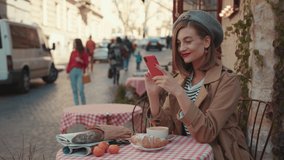 Smiling young woman with red lipstick hat wearing stylish coat typing on phone during sunny day sitting in cafe on breakfast outdoors city fashion online stylish slow motion