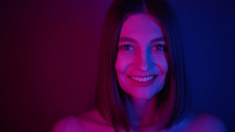 Wonderful young woman with bob hairstyle getting hair out of eyes, looking directly at camera. Smiling nice girl with dazzling smile. Purple neon background. Colorful.