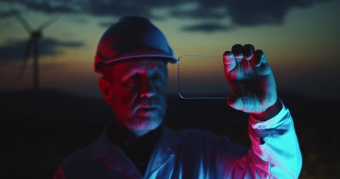 Portrait of focused male technician in uniform using mock-up transparent smartphone display working on wind turbines station at night.