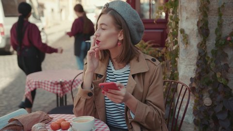 Smiling young blonde girl woman smoking cigarette at cafe smile wearing stylish coat use phone sunny day sitting in cafe on breakfast croissant morning outdoors smile city fashion online slow motion