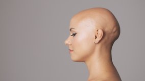 Side view of nice young hairless woman with fashion eye makeup runs a hand along the profile of the head isolated over gray wall
