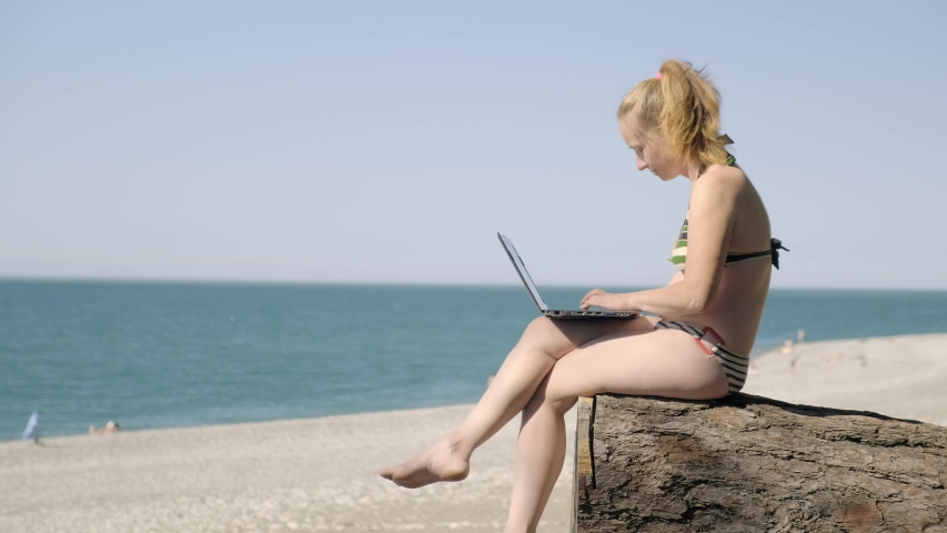Woman freelancer working on laptop while sitting on sea beach on log.