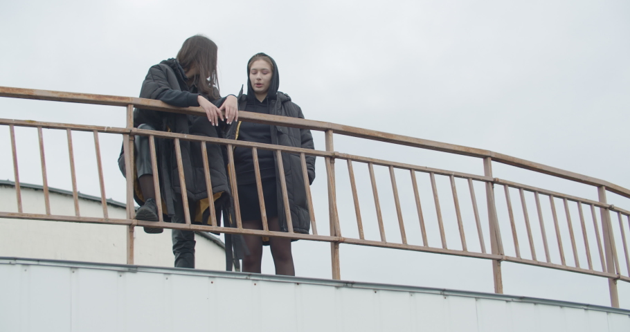 Two young attractive women wearing black coat with hood standing at rooftop of parking at city center. Pretty naughty arabian and caucasian fashion girls friends