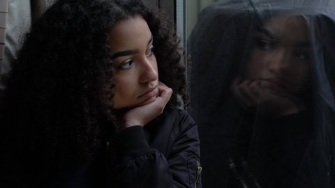 Sad depressed beautiful female mixed race African American girl teenager young woman teen leaning on her hand sitting looking out of a window in self isolation
