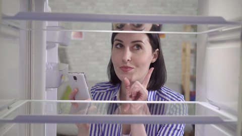 young woman looks into an empty fridge and orders food at home through an app in her phone