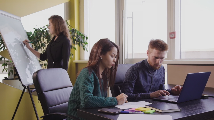 Group of american specialists work at table with laptop in ofice room. Businesspeople at desk. Beautiful woman coach write on whiteboard.