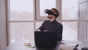 American businessman using vr glasses sitting at table with laptop in office. Front view of young man with black headset on face is watching video and playing game, get amazing virtual experience