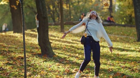 Young happy girl collecting fallen leaves throwing leaf rain swinging dancing enjoying life and sunshine in picturesque park. Fall season. Beautiful people.