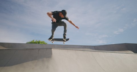 Skateboarder jumping over a concrete hip feature in a skatepark on a sunny day, slow motion skateboarding moves