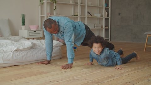Carefree affectionate black father and smiling positive preschool mixed race boy doing push-ups exercise in domestic room. Caring dad in casual clothes teaching little son doing push-ups at home. Stock Video