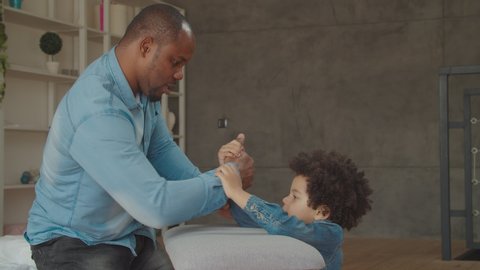 Positive diverse family with little kid competing in strength at home. Cheerful handsome african father and cute mixed race preschool son having fun, playing arm wrestling game during weekend.