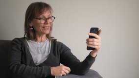 Happy middle aged mature woman holding smartphone having video chat, using social media apps in telephone sit on couch at home, older people and technology concept