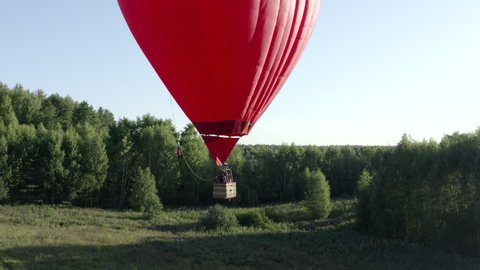 Hot air balloon in heart shape flying over green field and summer forest. Aerostat with basket flying while romantic trip in sky. Red love air balloon aerial view. 