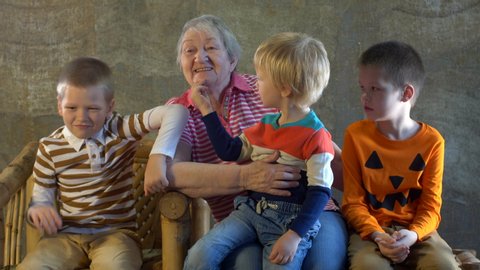Elderly female with gray hair, wrinkles and a beautiful face sits in front of the TV screen. Boys in bright clothes actively move and show emotions. Children love grandmother and hug granny
