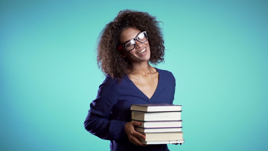 African student with glasses on blue background in the studio holds stack of university books from library. Girl smiles, she is happy to graduate.