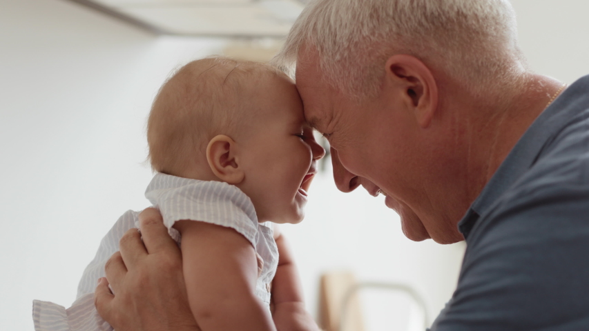 Funny Game and Laugh of Caucasian Old Man and Baby Girl in Domestic Comfort. Wrinkled Skin of Grandfather or Gray Haired Father in Casual Natural Lighting. Gentle Embrace and Happy Smile of 60s Parent | Shutterstock HD Video #1040853431
