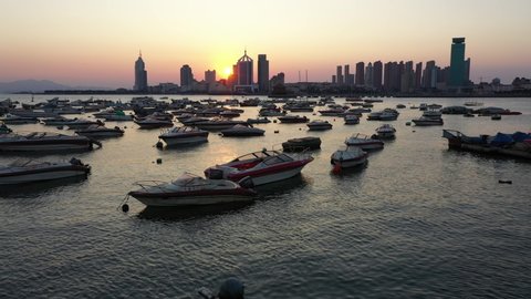 Flying over small fishing boats and recreational vessels towards modern city skyline of Qingdao at sunset, travel and tourism in China
