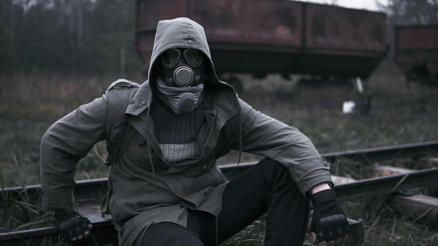 Man in gas mask and military clothes sitting on rail by abandoned rusty carriages at deserted train station. Gloomy landscape, post apocalypse, male wanderer survivor after nuclear disaster | Shutterstock HD Video #1040861855