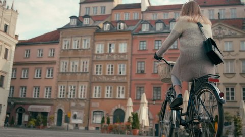 Pretty Woman in Coat Rides Bicycle in City Square with Old Buildings in Autumn Day. Urban Cycle Chic and European Architecture. 4K Slow motion Low Angle Tracking Shot with Copy Space