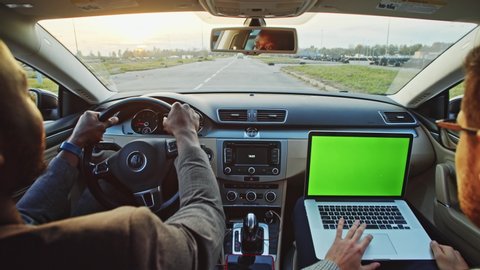 Afro-american young businessman driving expensive modern car on country road. Inside his colleague or friend working on greenscreen mock-up laptop computer.