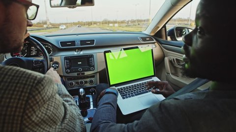 African man using modern laptop chrome key mock-up talking to colleague inside car. Couple of diverse friends driving car looking at green monitor laughing having fun.