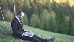 An Office Worker Crouched On The Grass In The Mountains And Is Typing Something On His Laptop. He Escaped From The City Noise To Rest. Business Concept Video