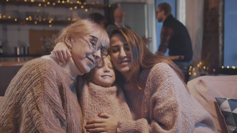 Portrait of three multi-aged beautiful women hugging and kissing on couch. Large family gathering together to celebrate christmas in modern decorated apartment.