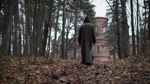 Mysterious monk or wizard in hooded robe walks throw autumn forest to medieval castle or monastery. Rear view. - Βίντεο στοκ