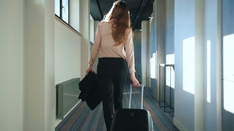 Businesswoman with travel suitcase walking at hotel corridor. Traveling woman with baggage settling in business hotel. Woman arriving in hotel for business trip