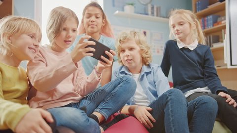 Kids Browsing on Internet and Playing Online Video Games on Mobile Phone, Watching Videos. Diverse Group of Cute Small Children Sitting together on the Bean Bags Use Smartphone and Talk, Have Fun.