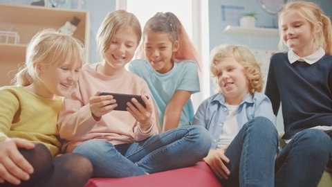 Kids Browsing on Internet and Playing Online Video Games on Mobile Phone, Watching Videos. Diverse Group of Cute Small Children Sitting together on the Bean Bags Use Smartphone and Talk, Have Fun