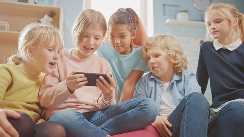 Diverse Group of Cute Small Children Sitting together on the Bean Bags Use Smartphone and Talk, Have Fun. Kids Browsing on Internet and Playing Online Video Games on Mobile Phone, Watching Videos