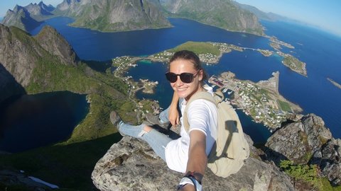 Young woman video blogger taking a selfie at the edge of a cliff with a top view to the Reine, Lofoten Islands, Norway.