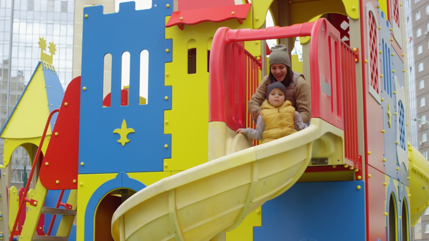 Tracking shot of young Caucasian mom sitting on top of playground slide and launching down son, boy descending on stomach, cheerful dad catching him at bottom, lifting up in air and spinning around