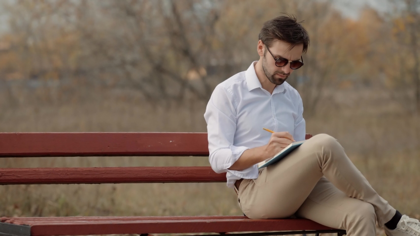 Man Sitting On Bench. Businessman Relaxing. Freelancer Drawing Sketch With Pencil Sitting On Bench. Male Hand Writing With Pen On Dairy. Businessman Working On Report And Writing Memories In Notebook