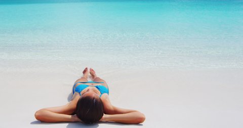 SEAMLESS LOOP VIDEO: Beach travel woman sunbathing relaxing lying down at perfect paradise beach. Relaxed woman in blue bikini lying on beach on summer vacation. Idyllic beach at luxury island resort.