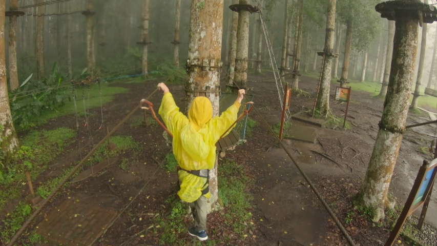 Action camera shot of a little boy in a safety harness climbs on a route in treetops in a forest adventure park. He climbs on high rope trail. Outdoor amusement center with climbing activities