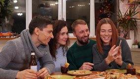 Young lady shows to her friends photos, videos from the smartphone. Happy mates are watching on phone, talking at festive table and celebrating new years eve party at home. Holidays concept 4K footage