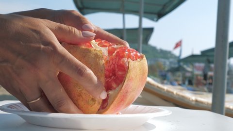 Female hands opens and shares a pomegranate close up. Woman preparing to eat juicy garnet at the beach. Hot summer day. Healthy food and veganism concept.