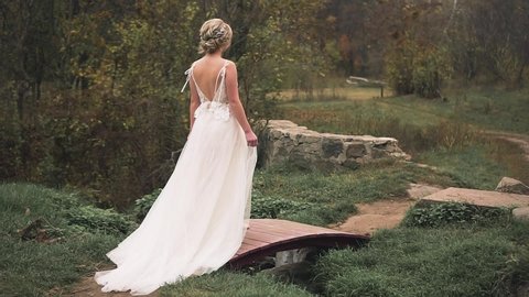 young blonde woman with collected elegant fashionable hairstyle walks in nature. white long vintage dress with train and open back. Shooting without face, cute bride looks around. Glamor girl princess