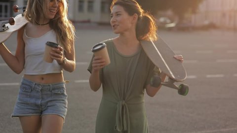 portrait of two girls Teenage friends with skateboard in sunshine in street european town laughing at something outside and drink coffe. slow motion