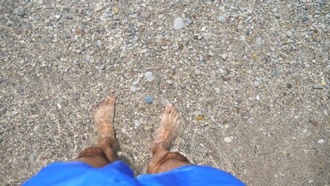 Ocean waves washing over male feet. Young man standing on seashore with pebble and enjoying tides of clear sea water. Guy enjoying summer vacation at seaside. Trip concept. Slow motion Top view POV