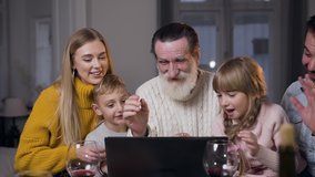 Charming smiling parents with their cute children and respected senior grandpa having video chat on laptop and saying hallo with waving hands to their computer friends