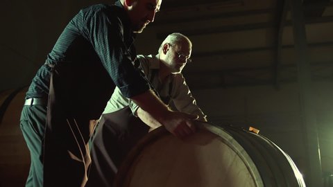 Worker in wine , whiskey or brandy warehouse sorting and rotating barrel . Two winemakers in vintage , traditional wine factory rolls barrel . Shot on ARRI ALEXA Cinema Camera in Slow Motion .
