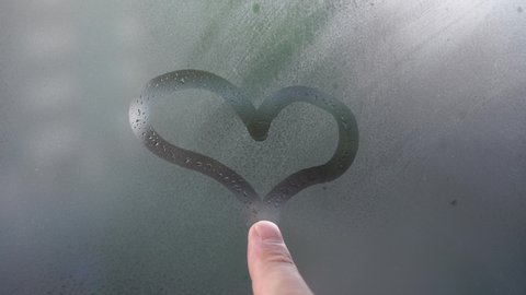 A finger draws a heart on a wet misted window. Romantic sign on glass in rainy weather. The concept of love and relationships