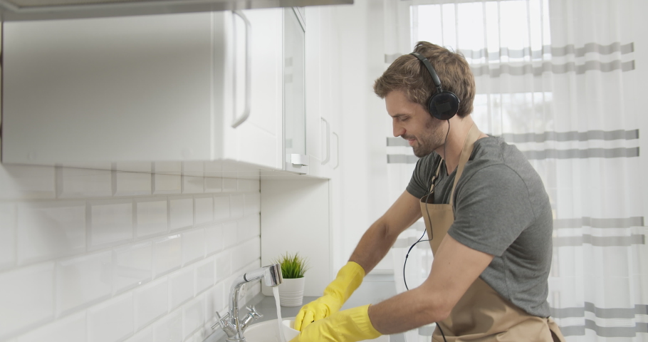 Handsome male in casual clothing and apron washing dishes, listening to favorite music in earphones and making dancing movements Royalty-Free Stock Footage #1041017243
