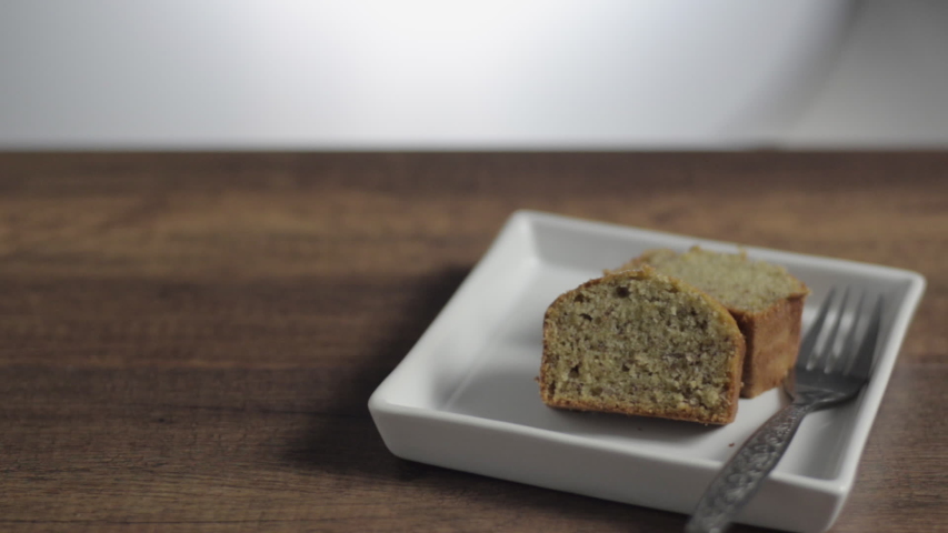 A hand putting a cup of tea down on a wood surface table with cut pieces of banana cake on a white plate.