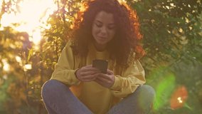 Pretty young woman with curly redhead hair using her smartphone while sitting in green park